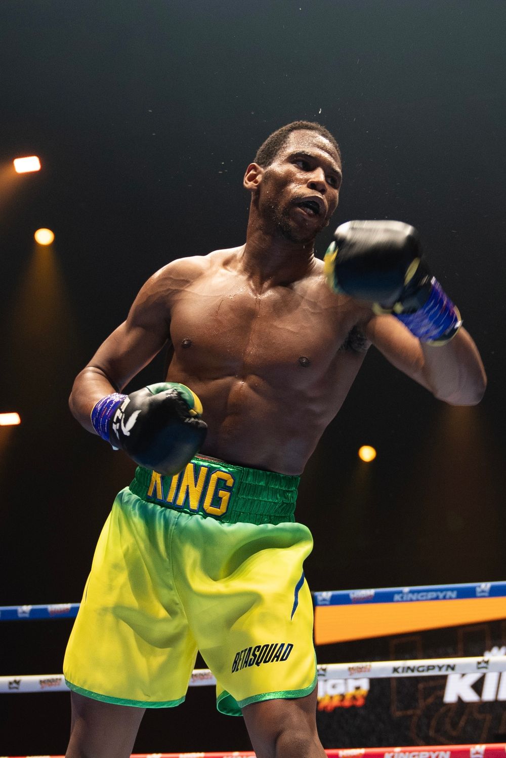 Muscular boxer in yellow and green King shorts competes under bright lights at a Kingpyn boxing event.