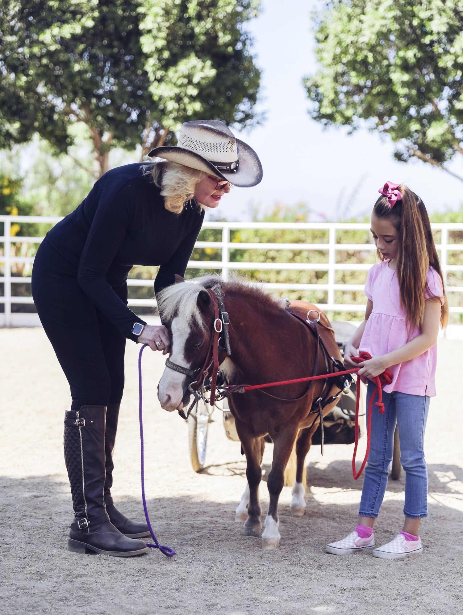 Woman in cowboy hat and boots teaching young girl to handle a small pony at an outdoor riding arena.