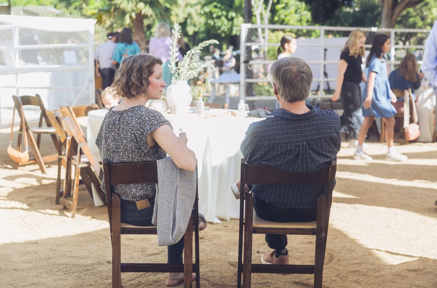 Couple seated at a white-draped table outdoors at a wedding reception venue with guests in the background.