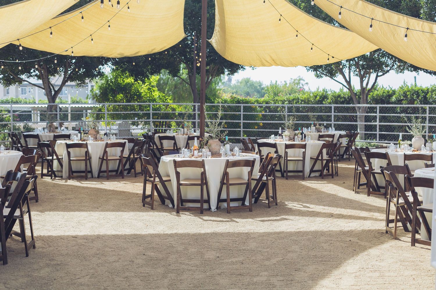 Outdoor wedding reception with round tables, white linens, dark wood folding chairs, string lights, and a yellow tent canopy.