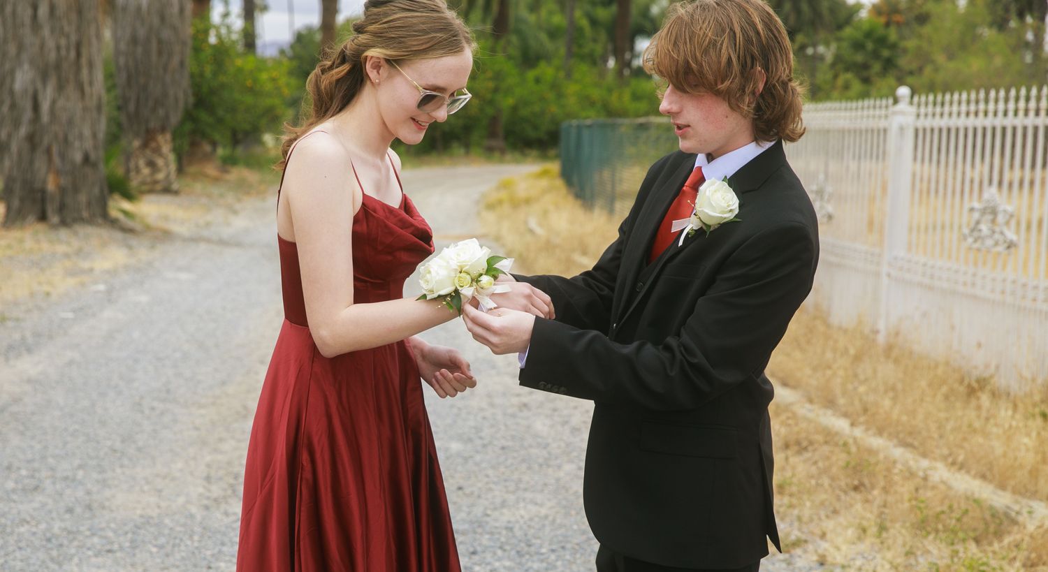A young couple in formal attire exchange a corsage outdoors, dressed for prom in a red gown and black tuxedo.