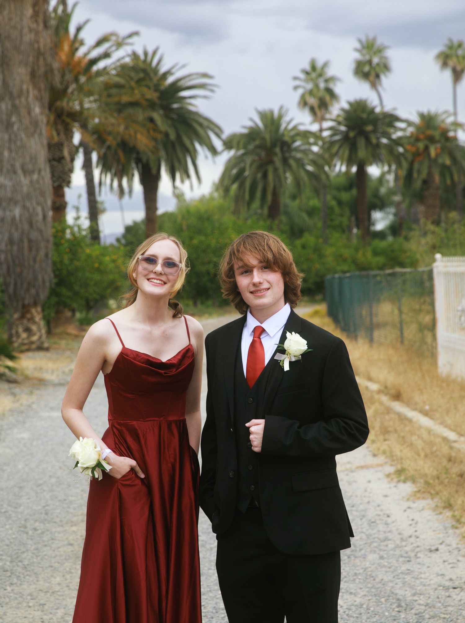 Teen couple in prom attire, girl in red gown with corsage, boy in black suit with red tie, posing outdoors.