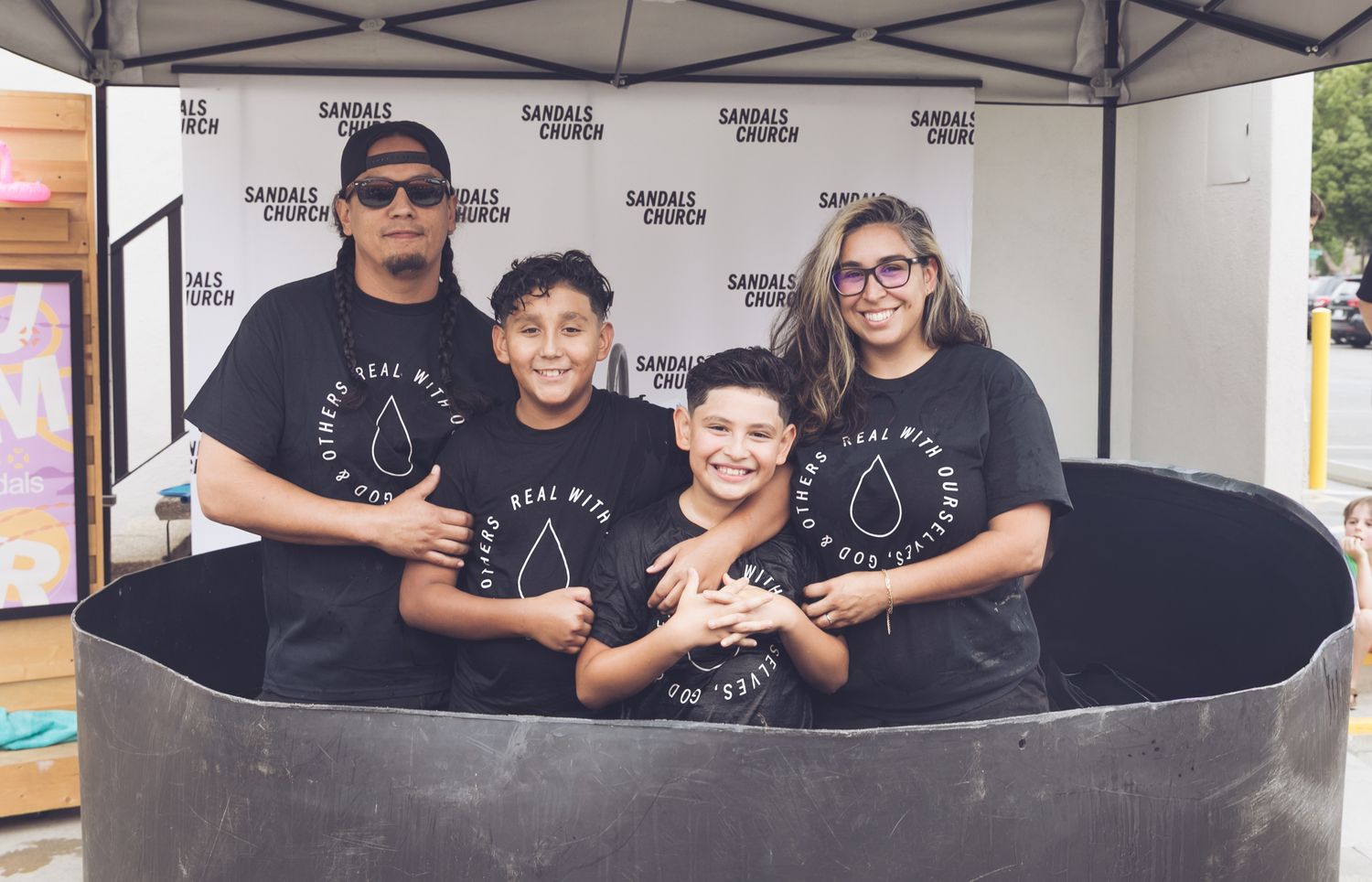 Family of four in matching black Sandals Church shirts posing together in a baptism pool at an outdoor church event.