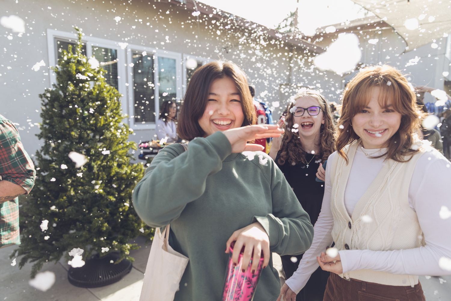 Three young women smile and laugh outdoors near a Christmas tree with artificial snow falling around them.