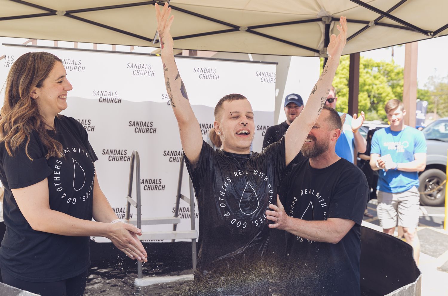 Joyful baptism ceremony outdoors at Sandals Church with three people in black shirts celebrating under a tent.