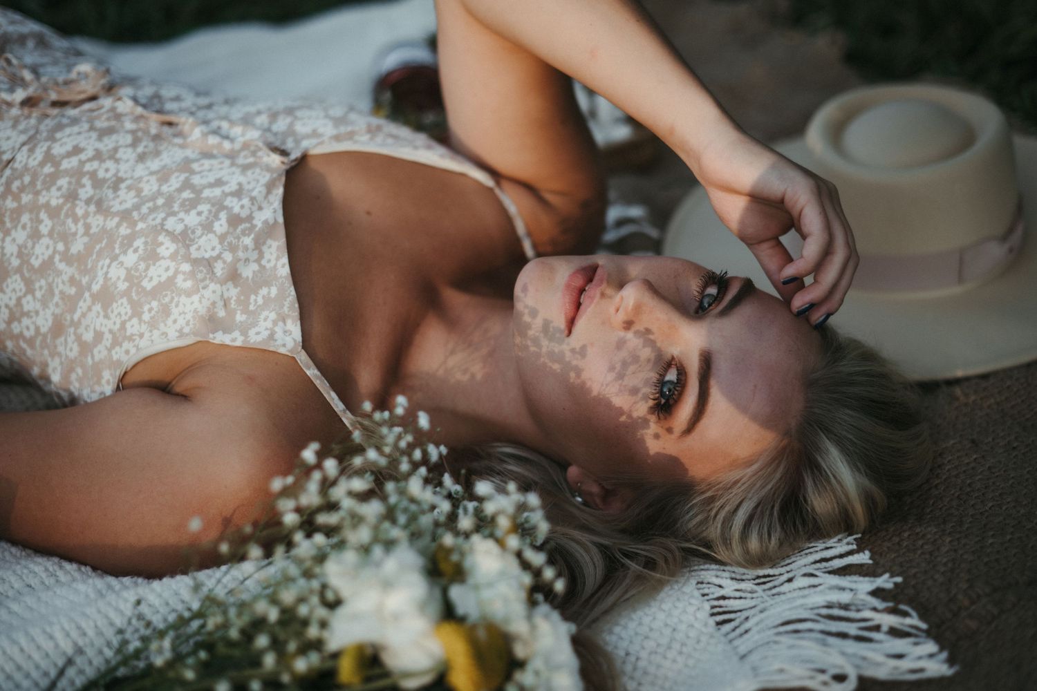 Blonde woman in lace dress lying on blanket with wildflower bouquet and straw hat in soft natural light in Knoxville, TN.