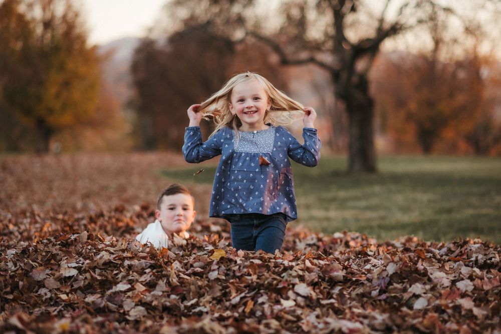 Two young children playing in a pile of autumn leaves outdoors, with a girl holding her hair up and smiling brightly in Johnson City, TN.