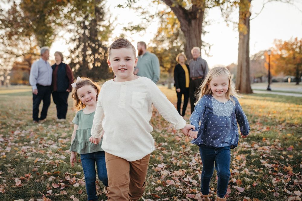 Three children hold hands running in a park during autumn, with family members standing among colorful fall trees in Johnson City, TN.