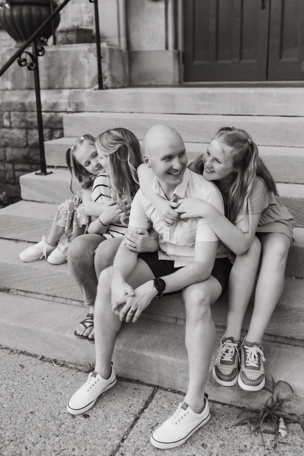 Black and white photo of a bald man sitting on steps laughing with two young girls hugging him in Louisville, KY.