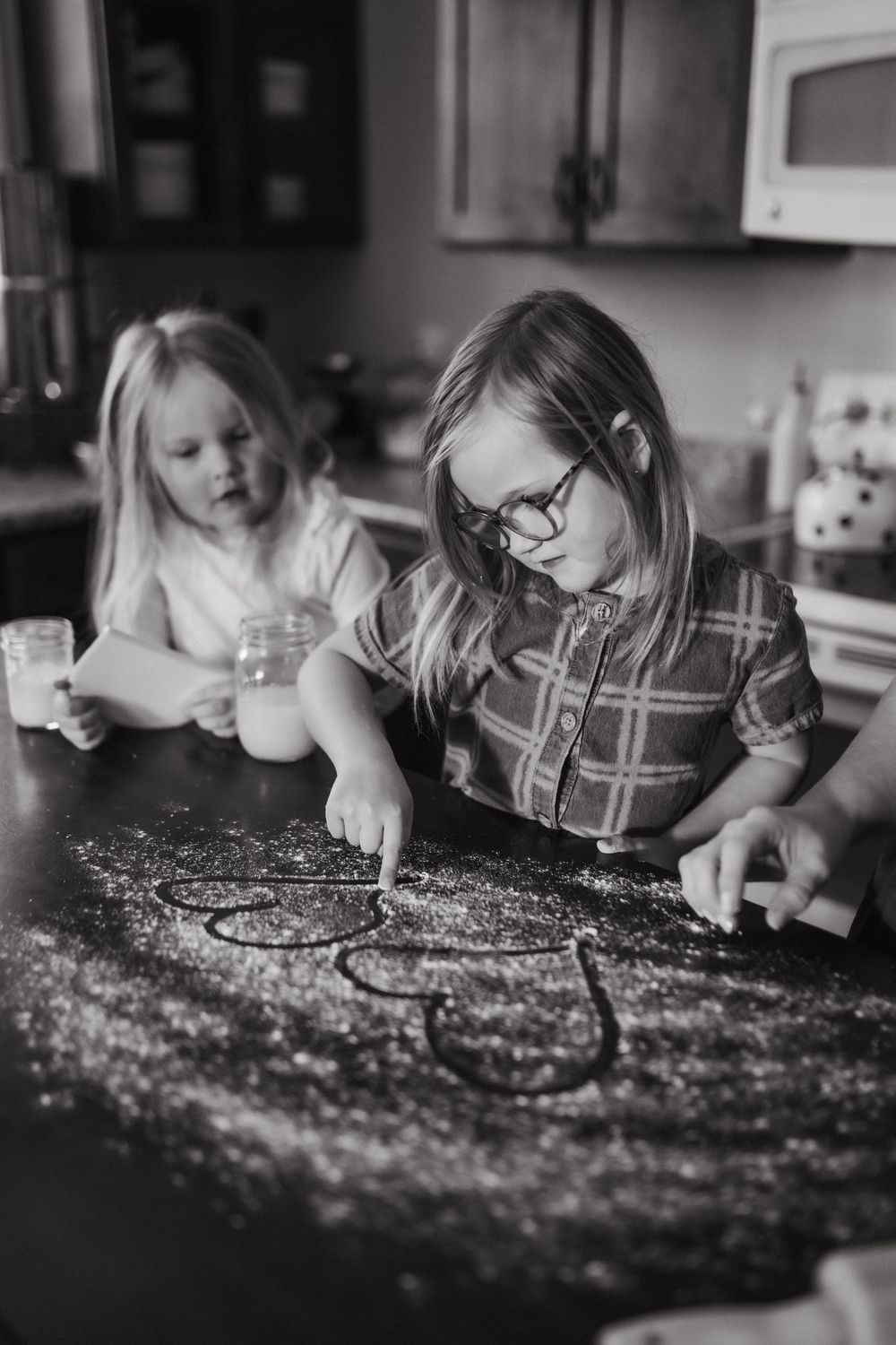 Two young girls drawing shapes in flour on a kitchen counter in a black and white photo in Johnson City, TN.