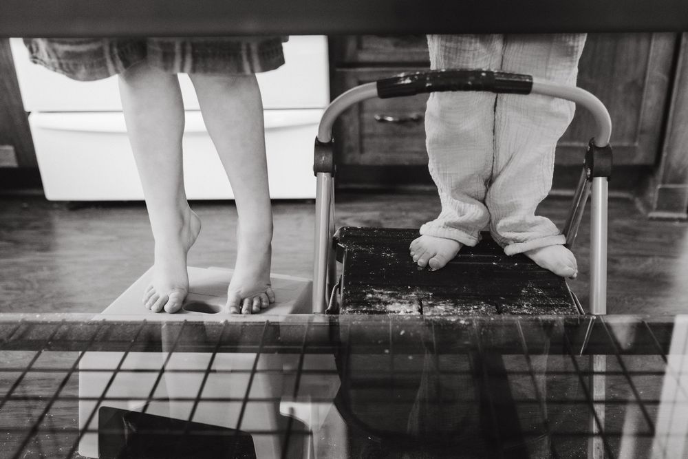 Black and white photo of bare feet and toddler in footie pajamas standing on chair under a table in Johnson City, TN.