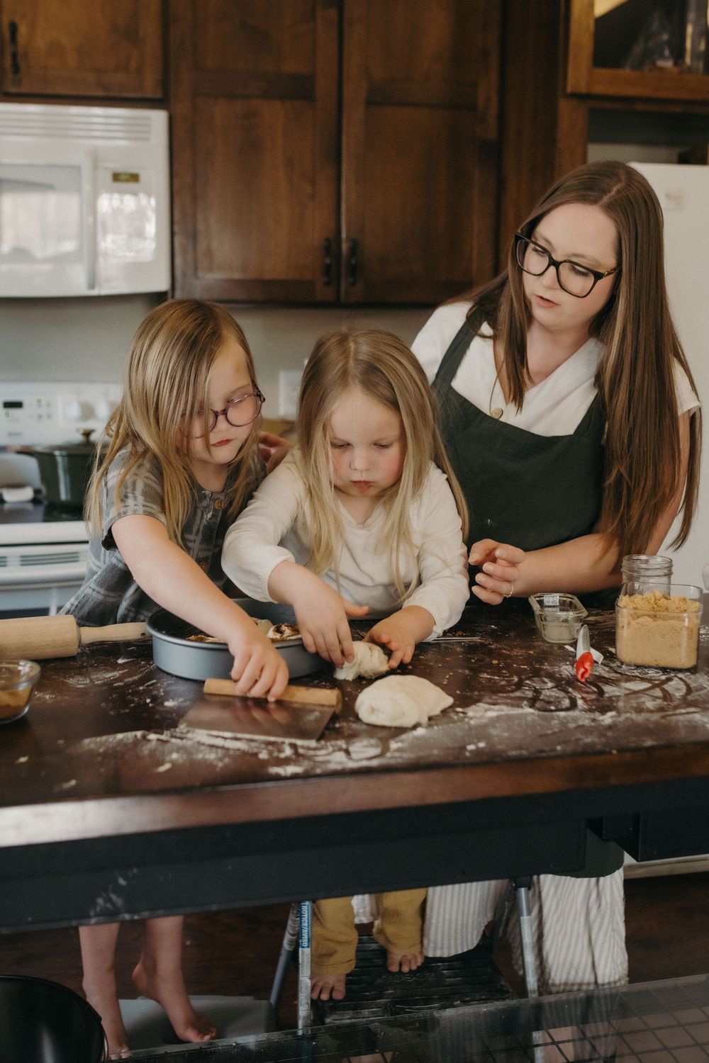 A mother and two young girls baking together in a rustic kitchen, rolling dough on a flour-dusted wooden counter in Johnson City, TN.