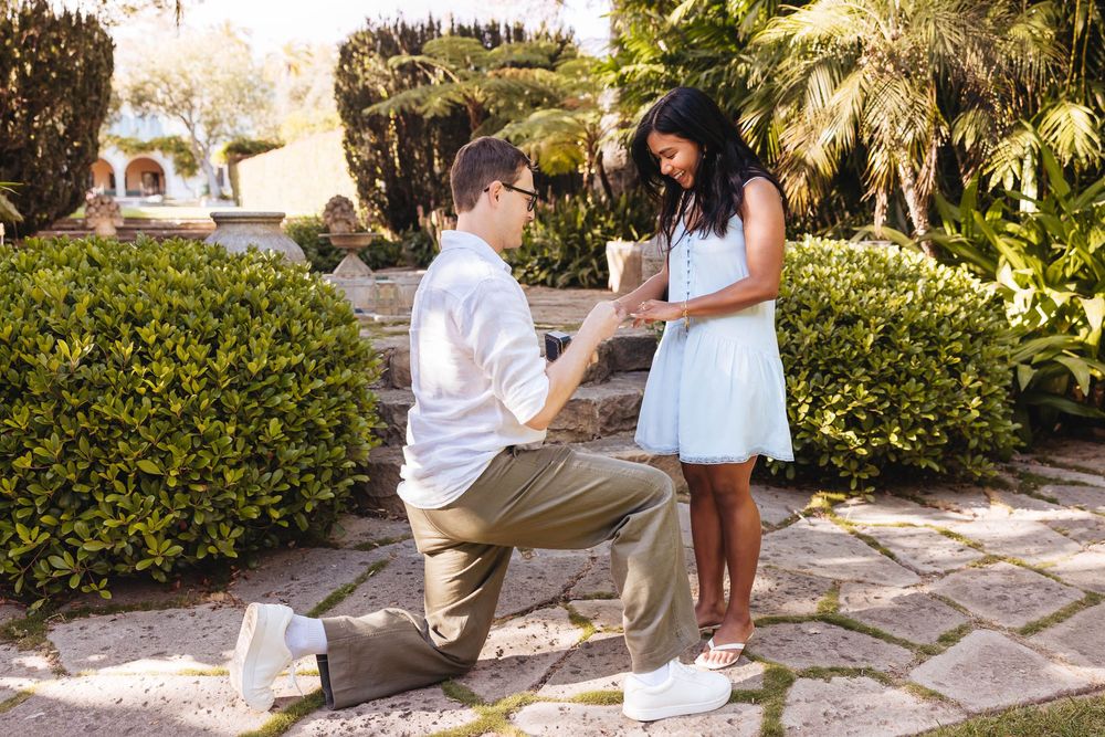 Man on one knee presenting engagement ring to woman in light blue dress in a garden setting.