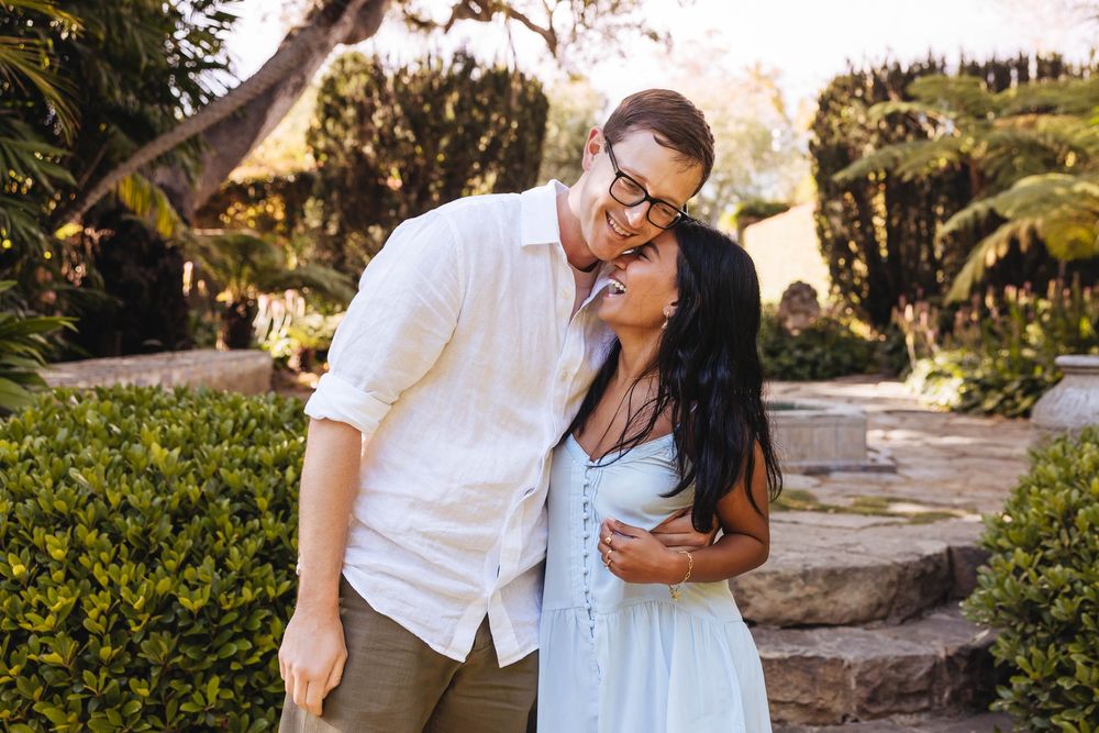 A couple embraces in a lush garden, the man in a white shirt gazing down at the woman in a light blue dress.