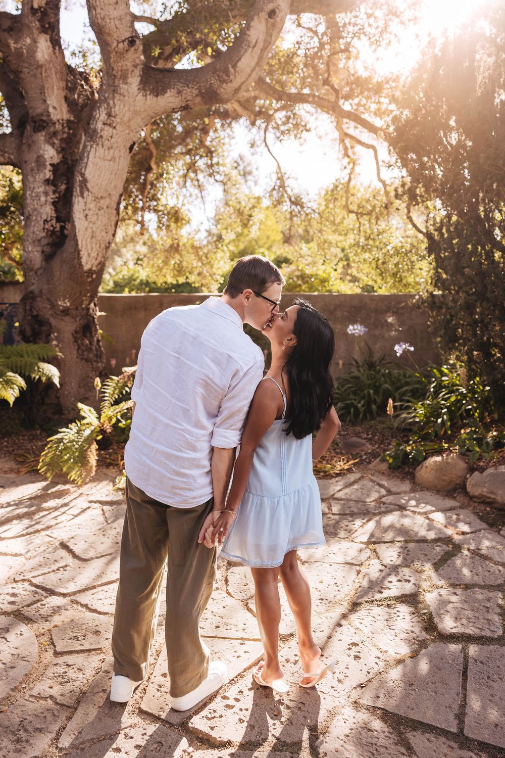 Couple sharing a romantic kiss in a sunlit garden with lush greenery and stone pathway.