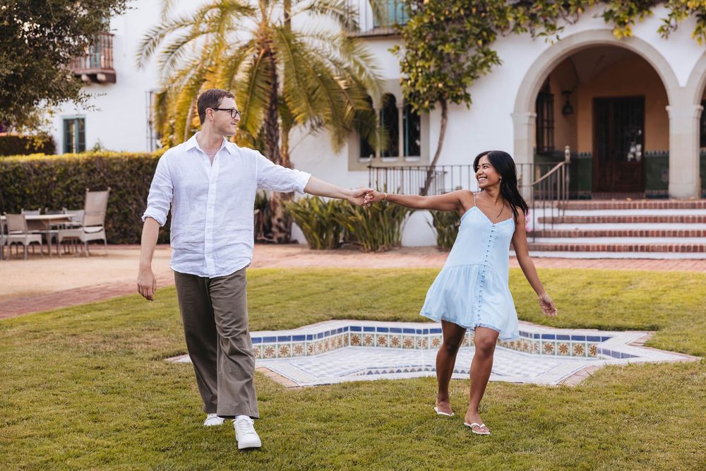 Couple dancing and holding hands on green lawn outside elegant white Spanish-style building with palm trees.