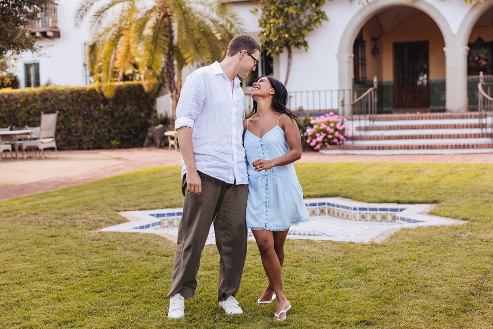 Couple sharing a romantic moment on lush green lawn outside elegant estate, woman in blue dress, man in white shirt.