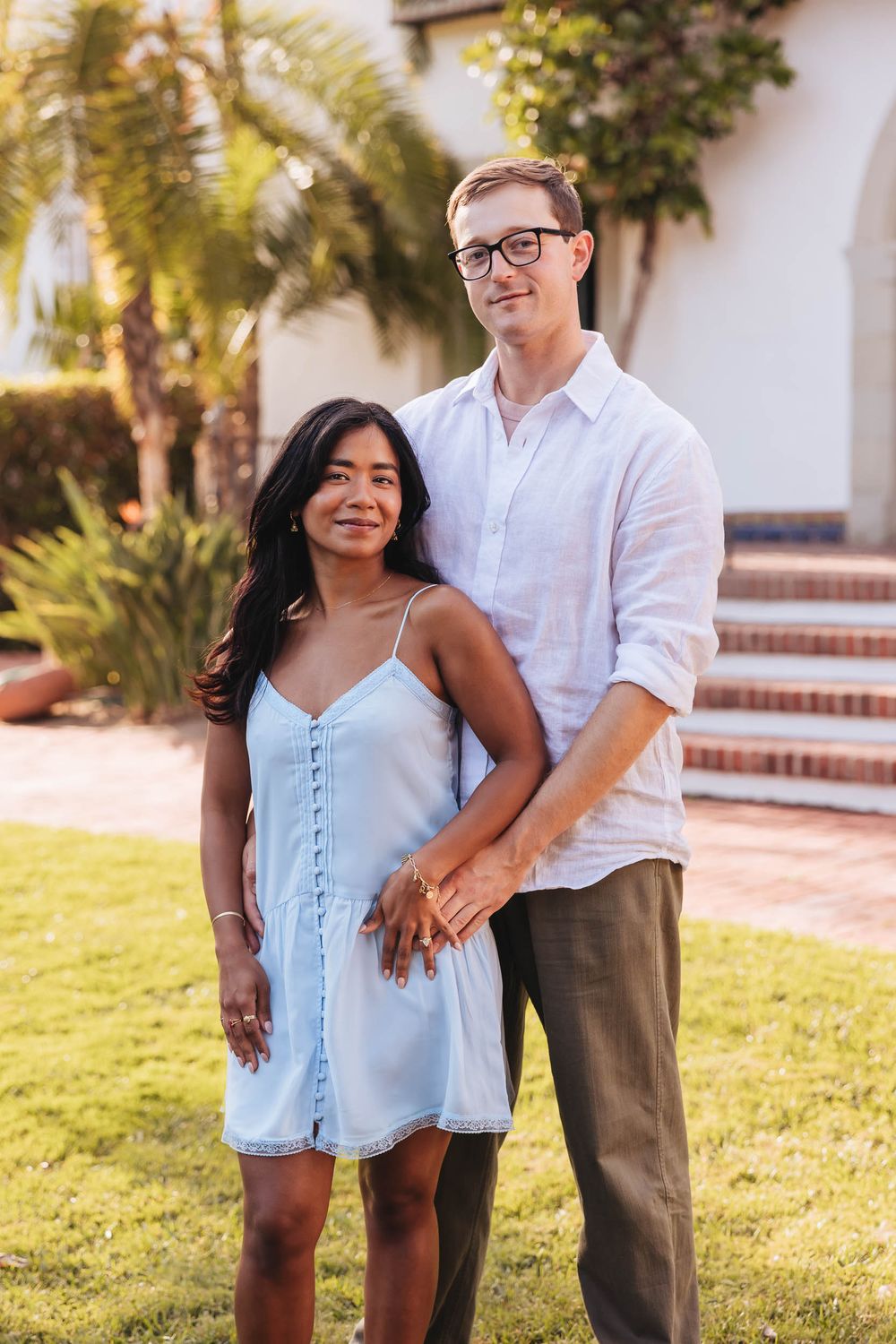 Couple holding hands smiling at camera outdoors near palm trees and brick steps in warm sunlight.
