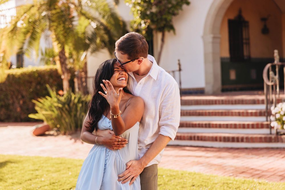 Couple embracing outdoors, woman showing engagement ring, smiling joyfully near brick steps and palm trees in warm sunlight.