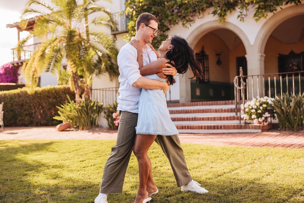 A joyful interracial couple dancing together on a green lawn outside a Mediterranean-style building.