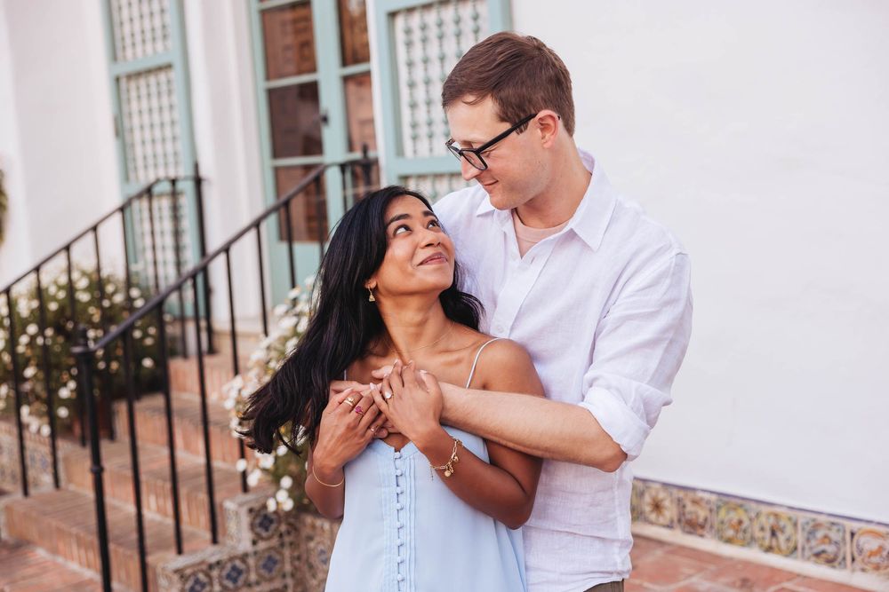A couple embraces outdoors, woman in blue dress gazes up at man in white shirt holding her hands near a building.