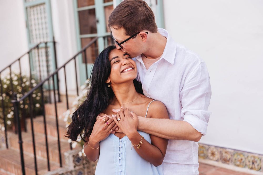 A happy interracial couple embracing outdoors, man kissing woman's cheek while holding her hands near a building.