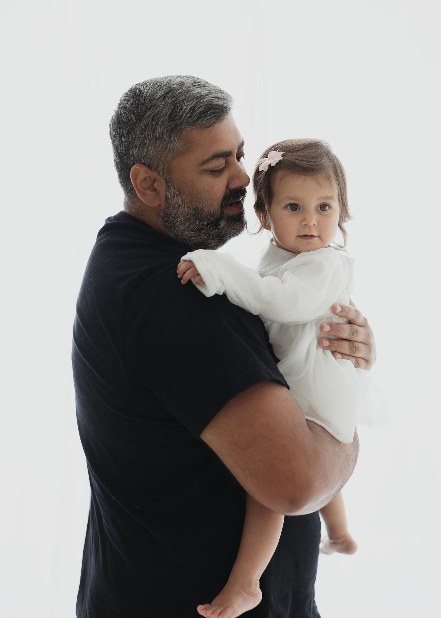 A bearded man in a black t-shirt tenderly holds a baby girl in a white outfit with a pink hair bow. Elizabeth Lang Photography Surrey