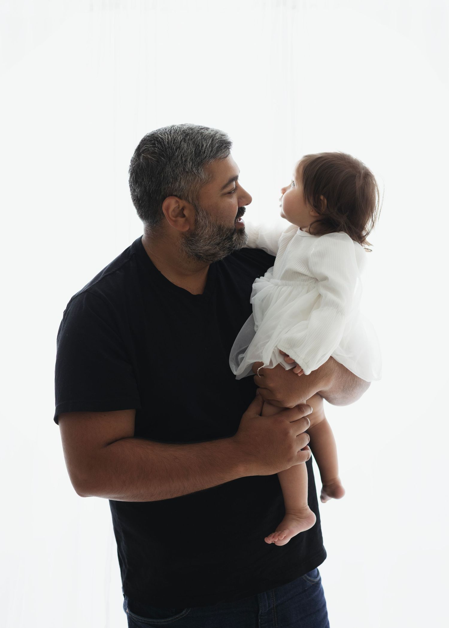 A man in a black t-shirt lovingly holds a baby in a white dress against a white background. Elizabeth Lang Photography Surrey