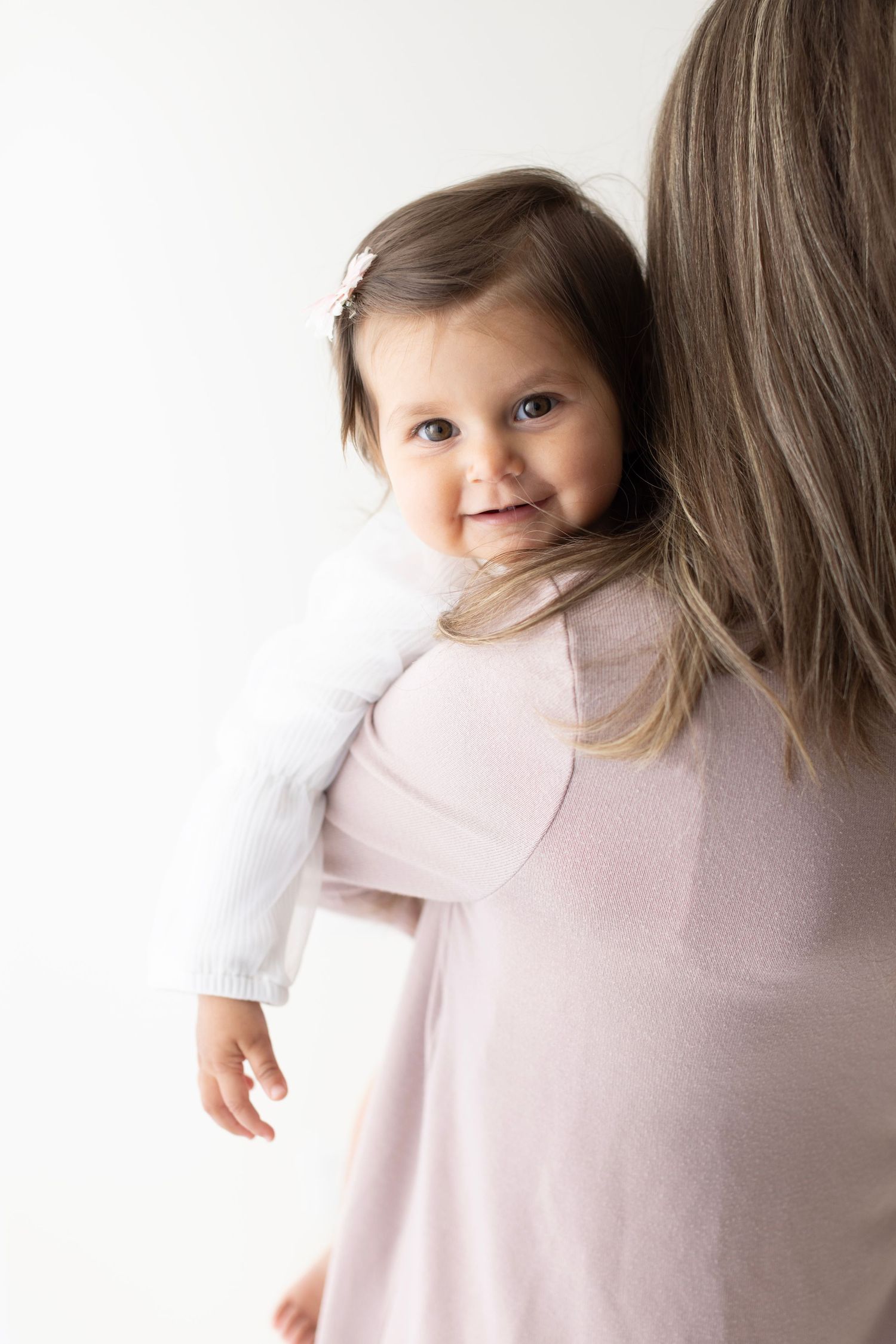 Smiling baby girl in white cardigan being held over shoulder of mother in pink dress against white background. Elizabeth Lang Photography Su