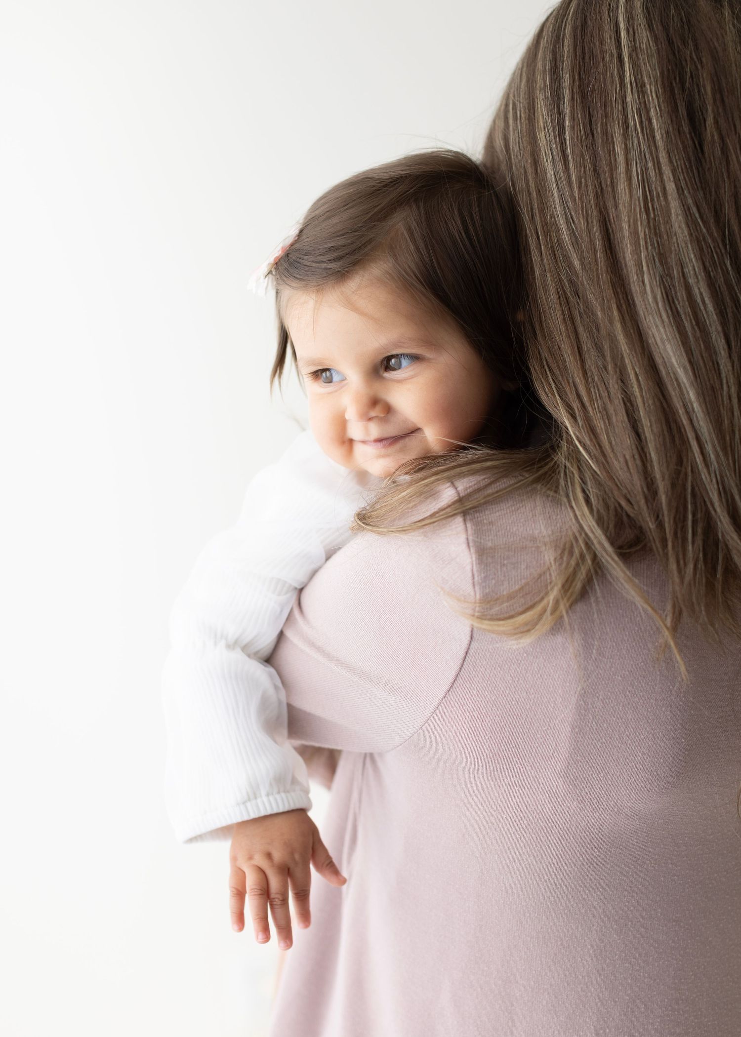 Adorable toddler girl with brown hair smiling over mother's shoulder wearing a white cardigan and pink dress. Elizabeth Lang Photography Sur