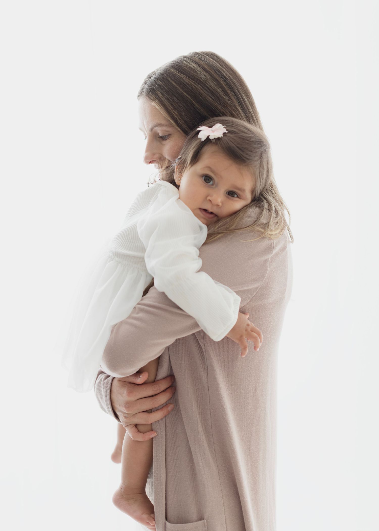 A mother in a pink dress tenderly holds her toddler girl in a white dress with a bow hair clip. Elizabeth Lang Photography Surrey