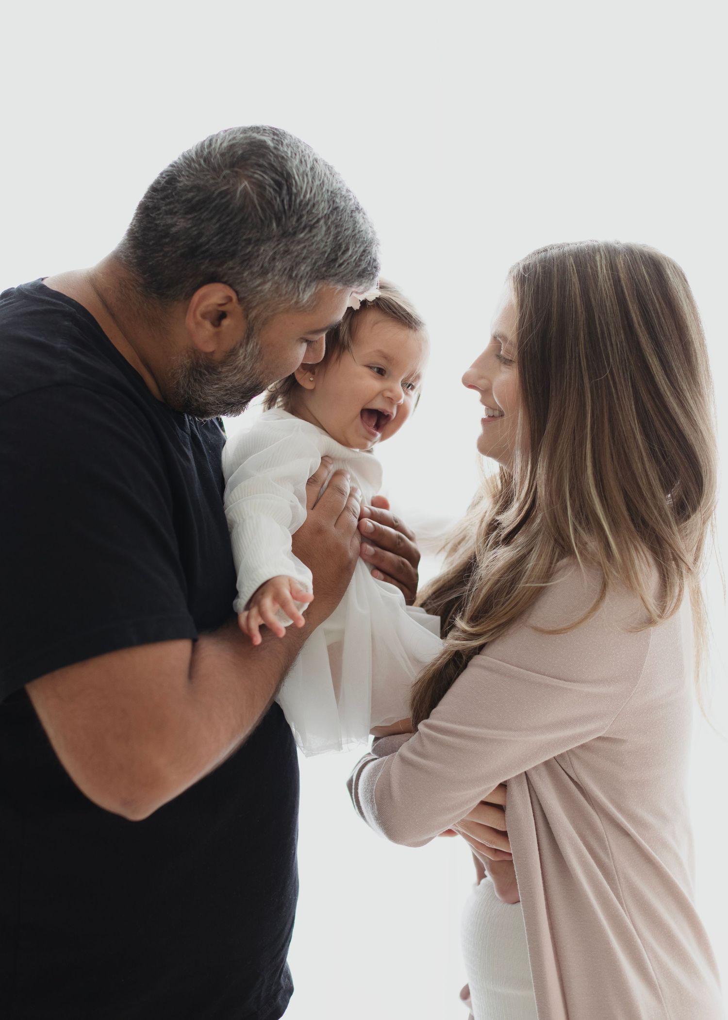 Happy baby laughing between loving parents during a bright family portrait session. Elizabeth Lang Photography Surrey