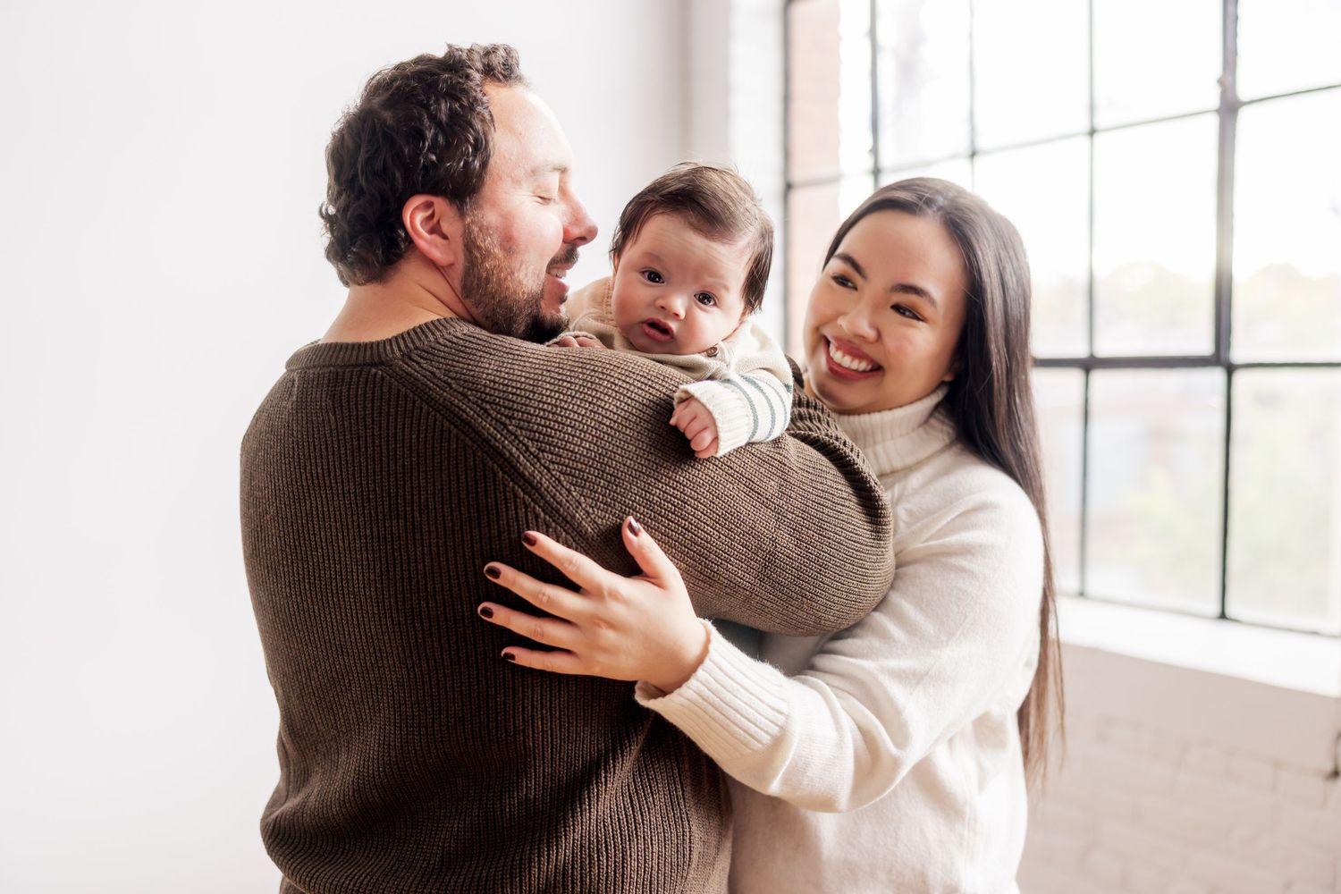 Happy young family with dark-haired mother, bearded father, and baby in denim outfit posing by large window.