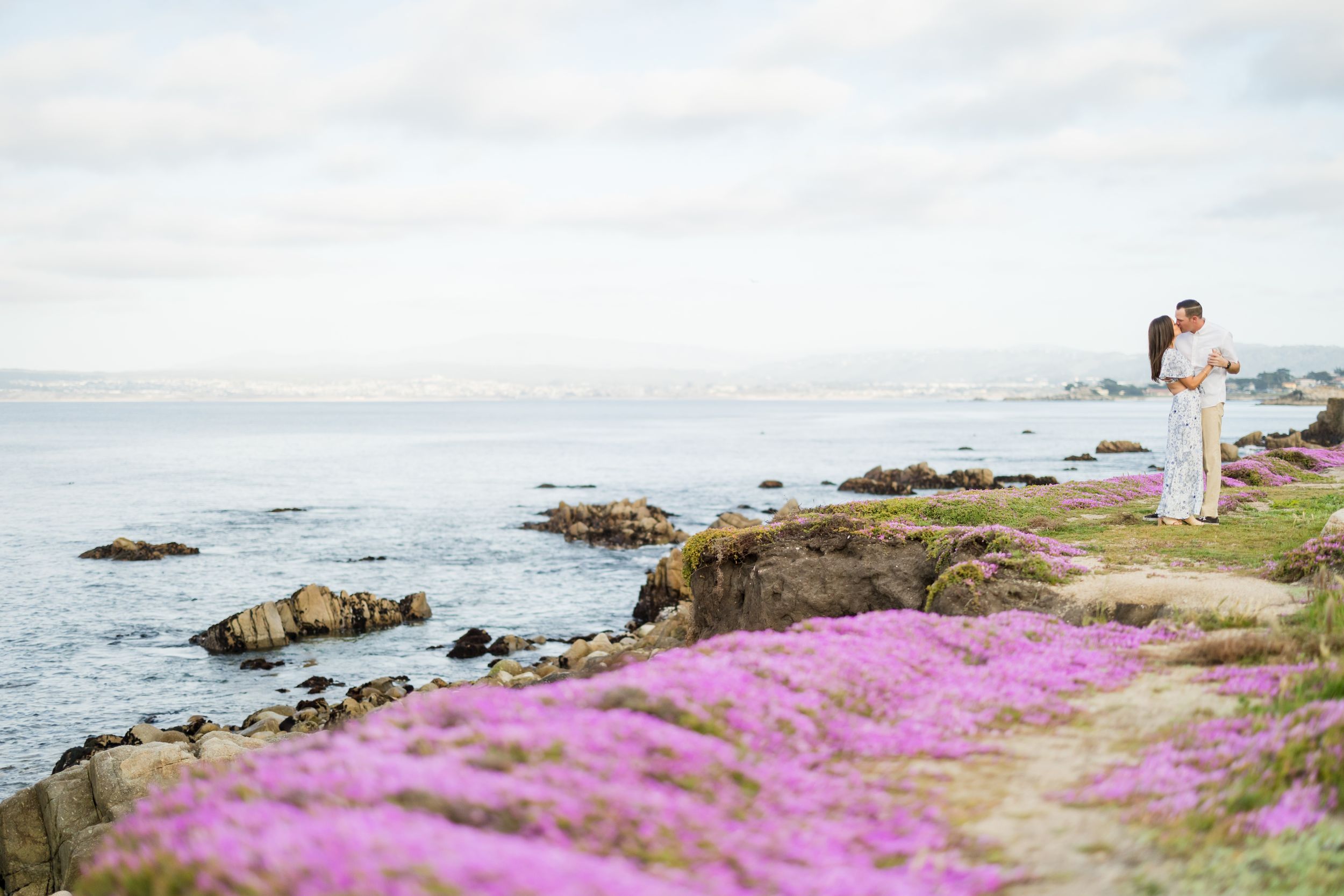 Couple embracing on rocky coastal cliffs surrounded by vibrant purple wildflowers with ocean views.