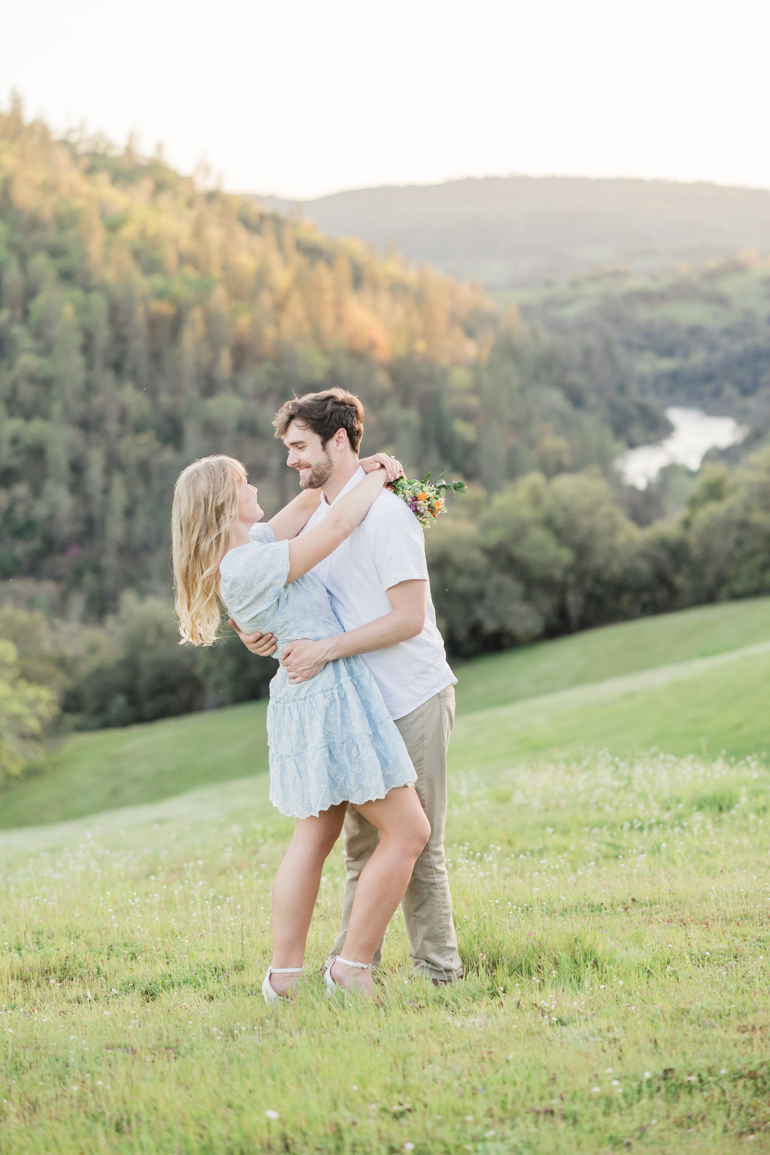 Couple embracing in a green meadow with rolling forested hills and a river at golden hour sunset.