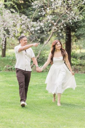 Couple holding hands walking on green grass near blooming cherry trees, woman in white dress, man pointing forward.