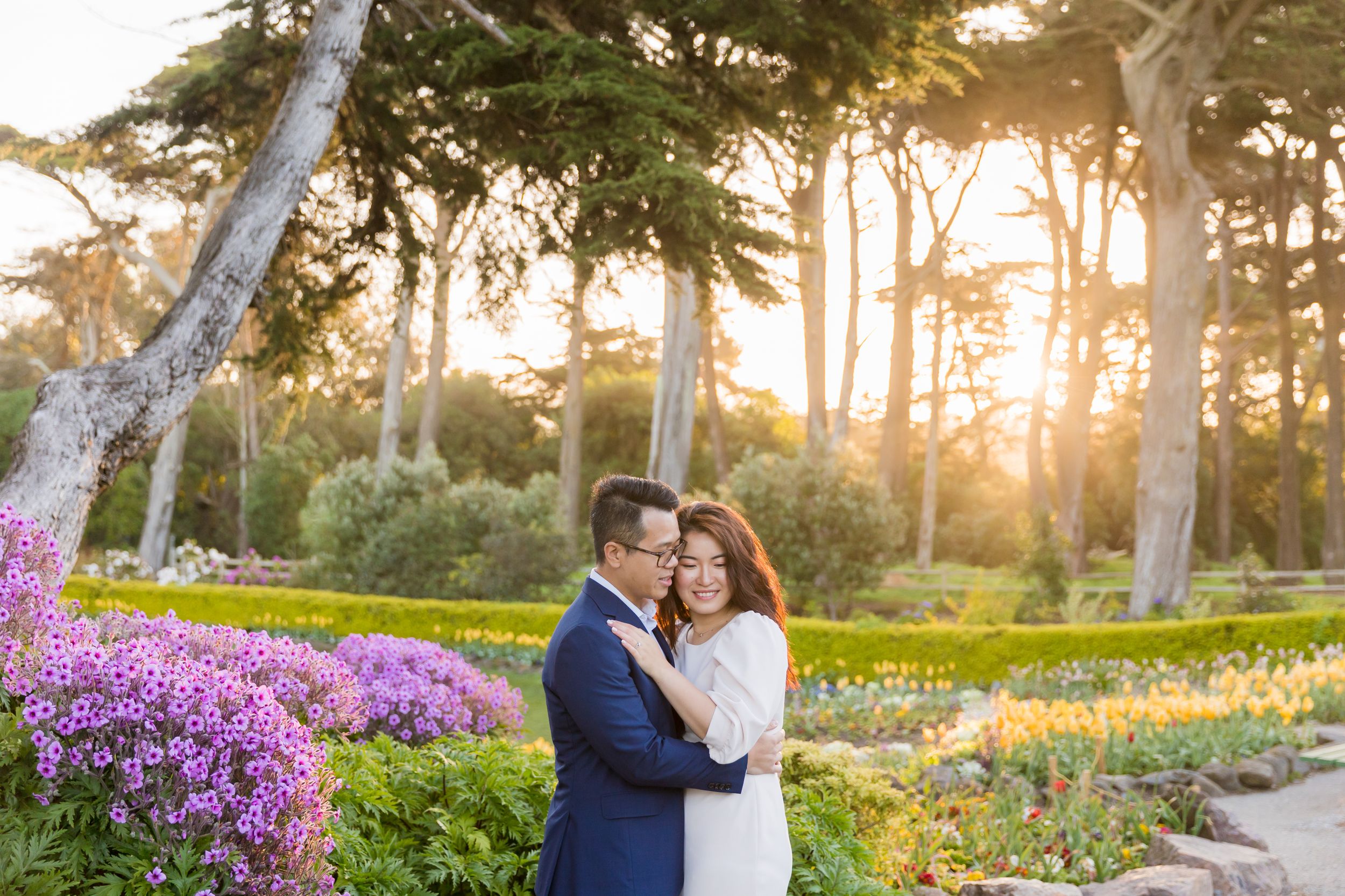 Couple embracing at sunset in a lush garden with purple flowers and tall trees in warm golden light.