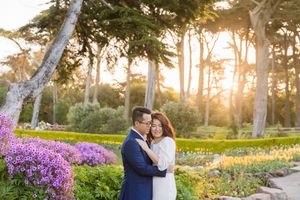 Couple embracing at sunset in a lush garden with purple flowers and tall trees in warm golden light.
