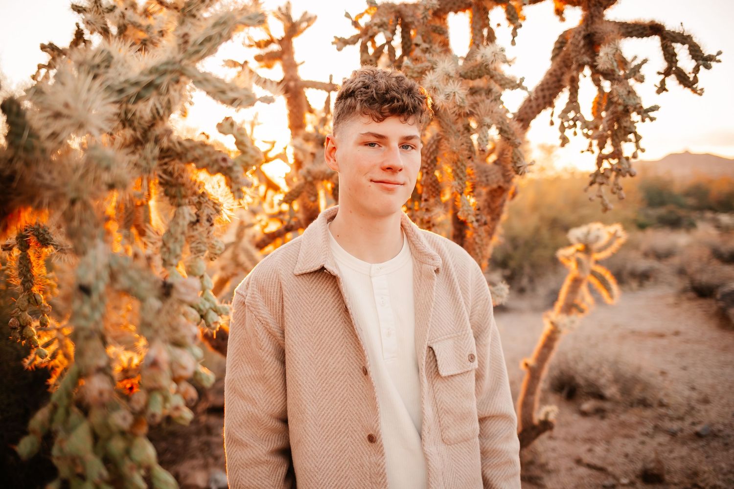 Young man in beige jacket standing before golden-lit cholla cactus in desert landscape at sunset.