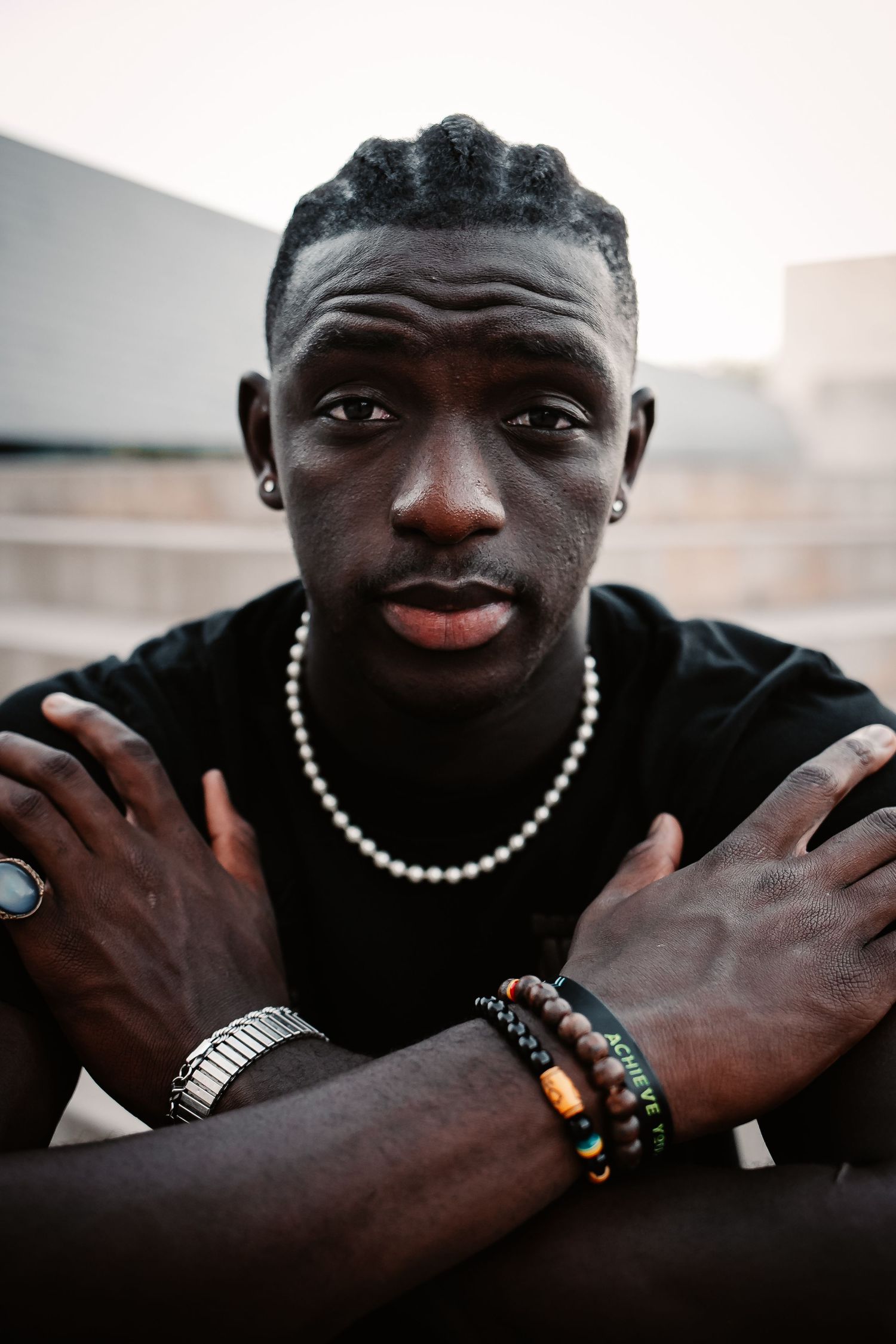 Young Black man in black t-shirt with pearl necklace and beaded bracelets, arms crossed, looking at camera outdoors.