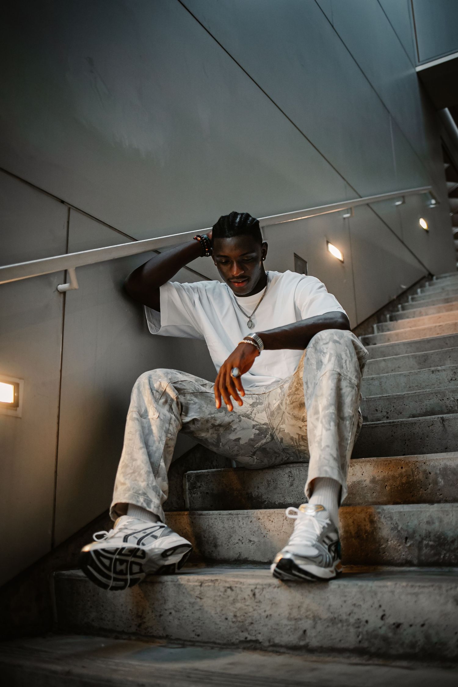 Young Black man in white tee and distressed pants sitting on concrete stairs, posing confidently with sneakers and chain necklace.