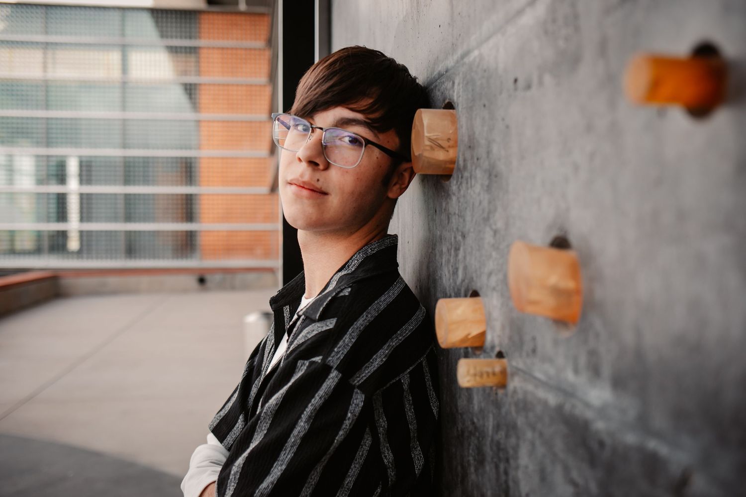 Young person with glasses leaning against a concrete wall outdoors, wearing a striped shirt.