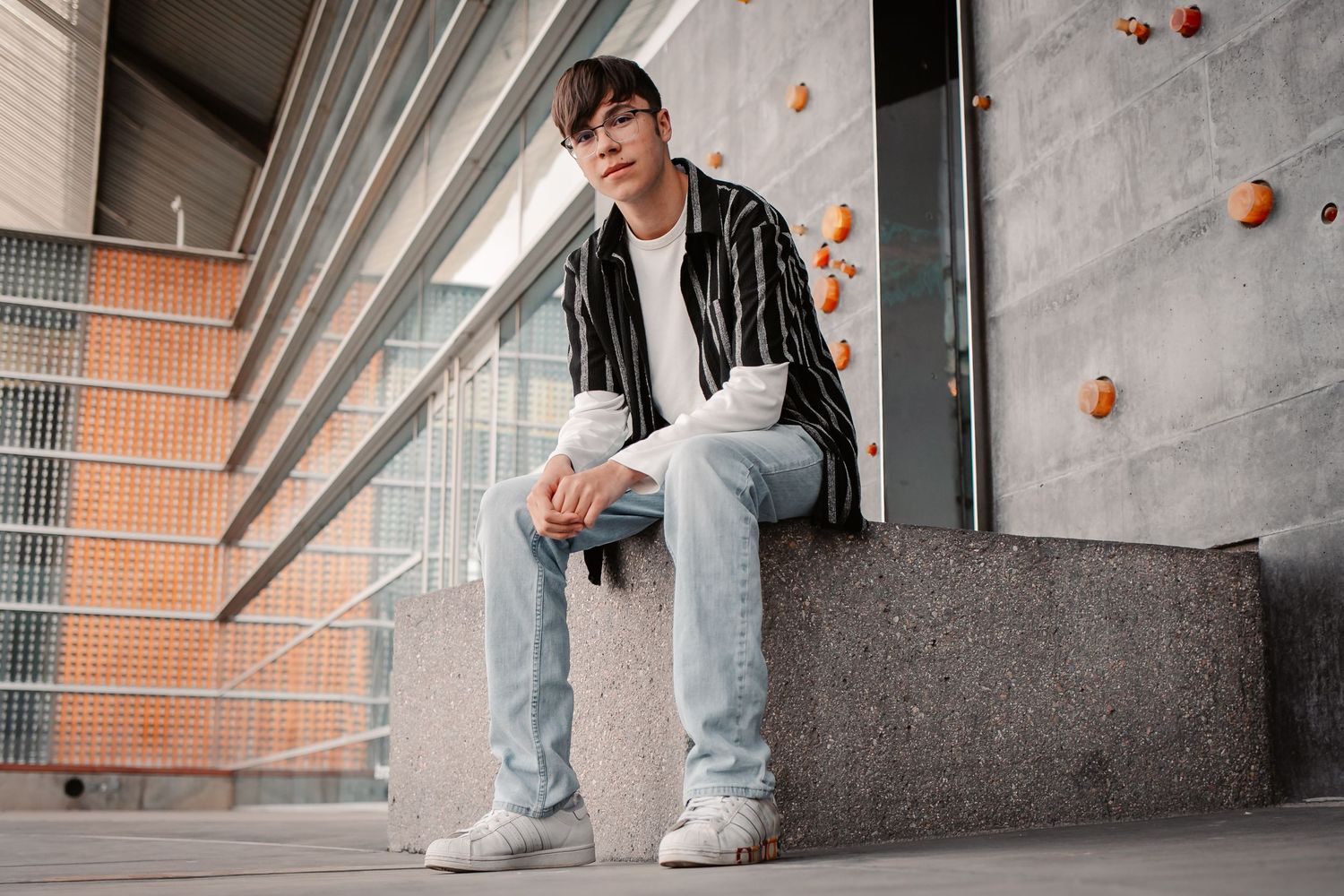 Young man in striped jacket and light jeans sitting on concrete ledge outside modern building.