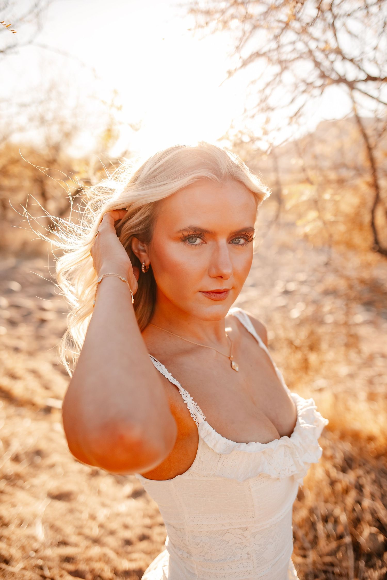 Blonde woman in white dress poses outdoors in golden sunset light among dry brush, hand in hair.