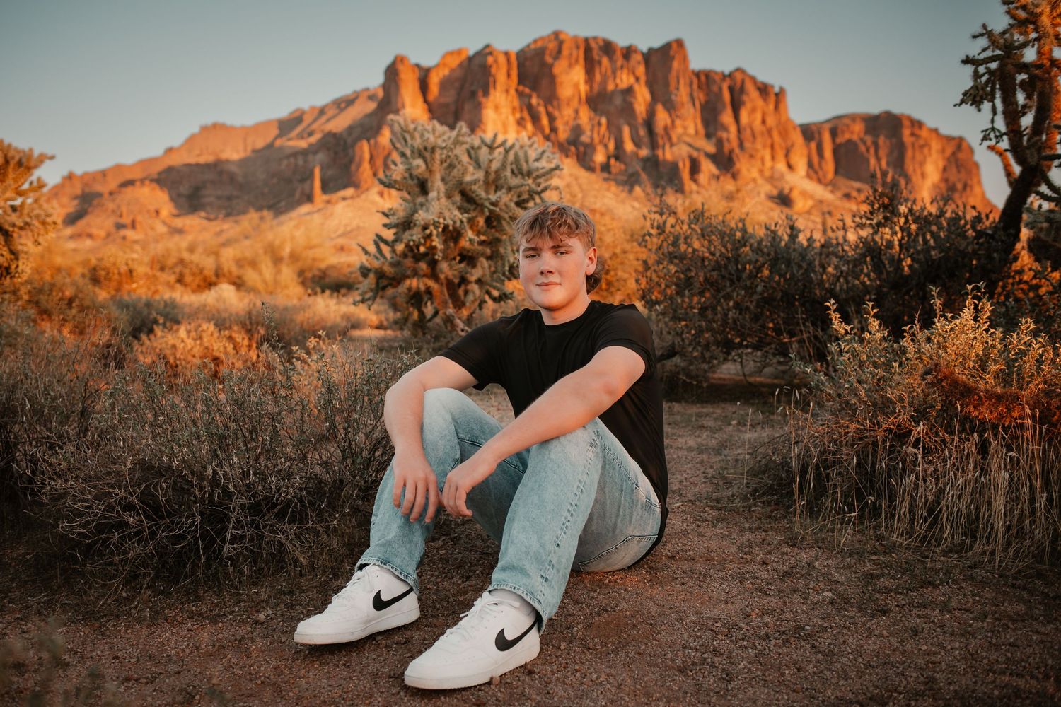 A young man in a black tee and jeans sits on desert ground before a sunlit rocky mountain at golden hour.
