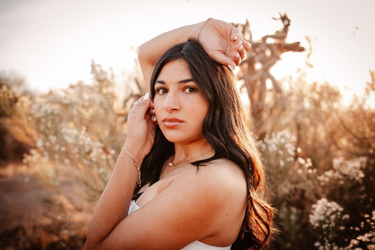 Young woman with long dark hair poses outdoors at golden hour among wild plants, wearing a white top and gold jewelry.