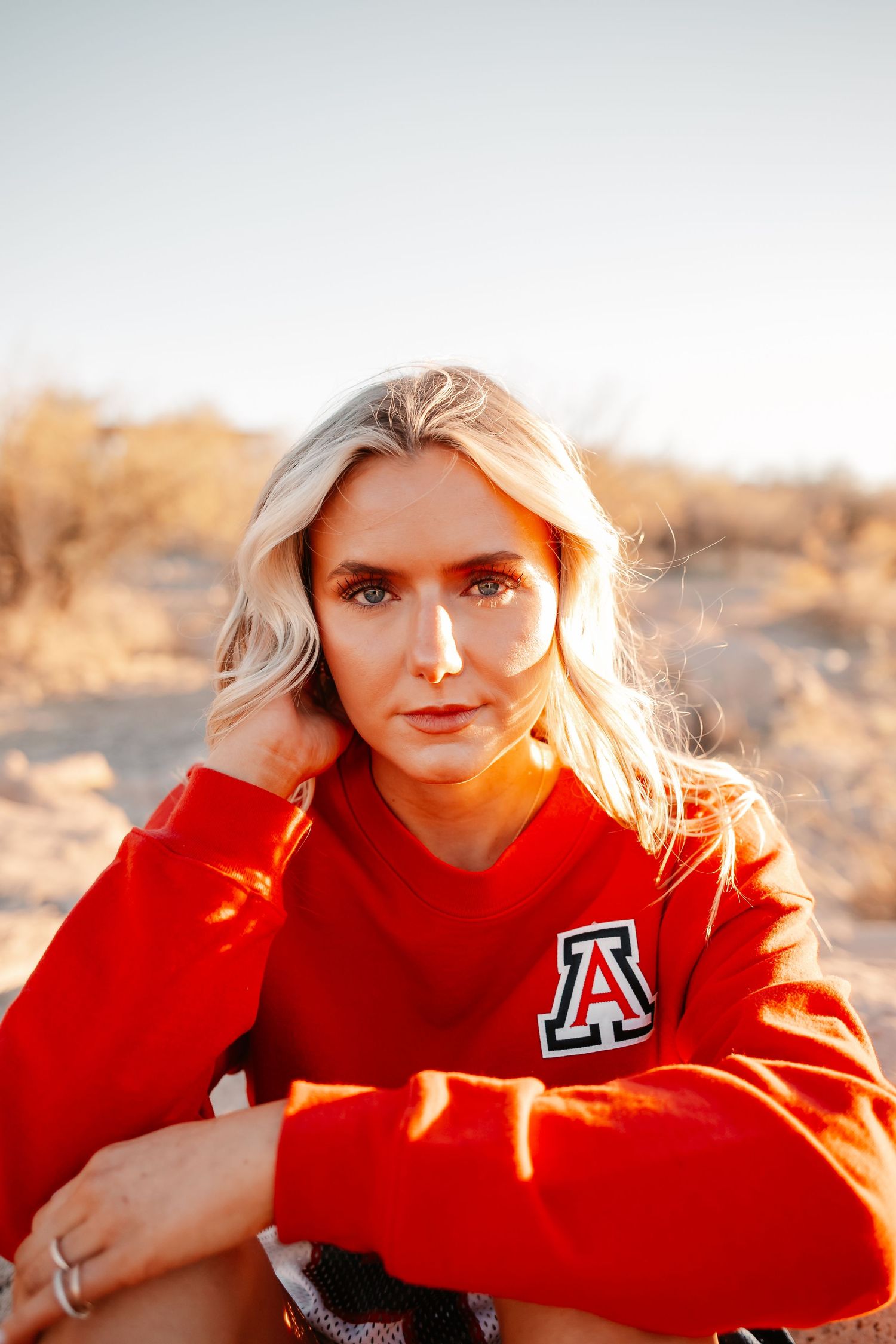 Blonde woman in red University of Arizona sweatshirt sitting outdoors in desert landscape at golden hour.