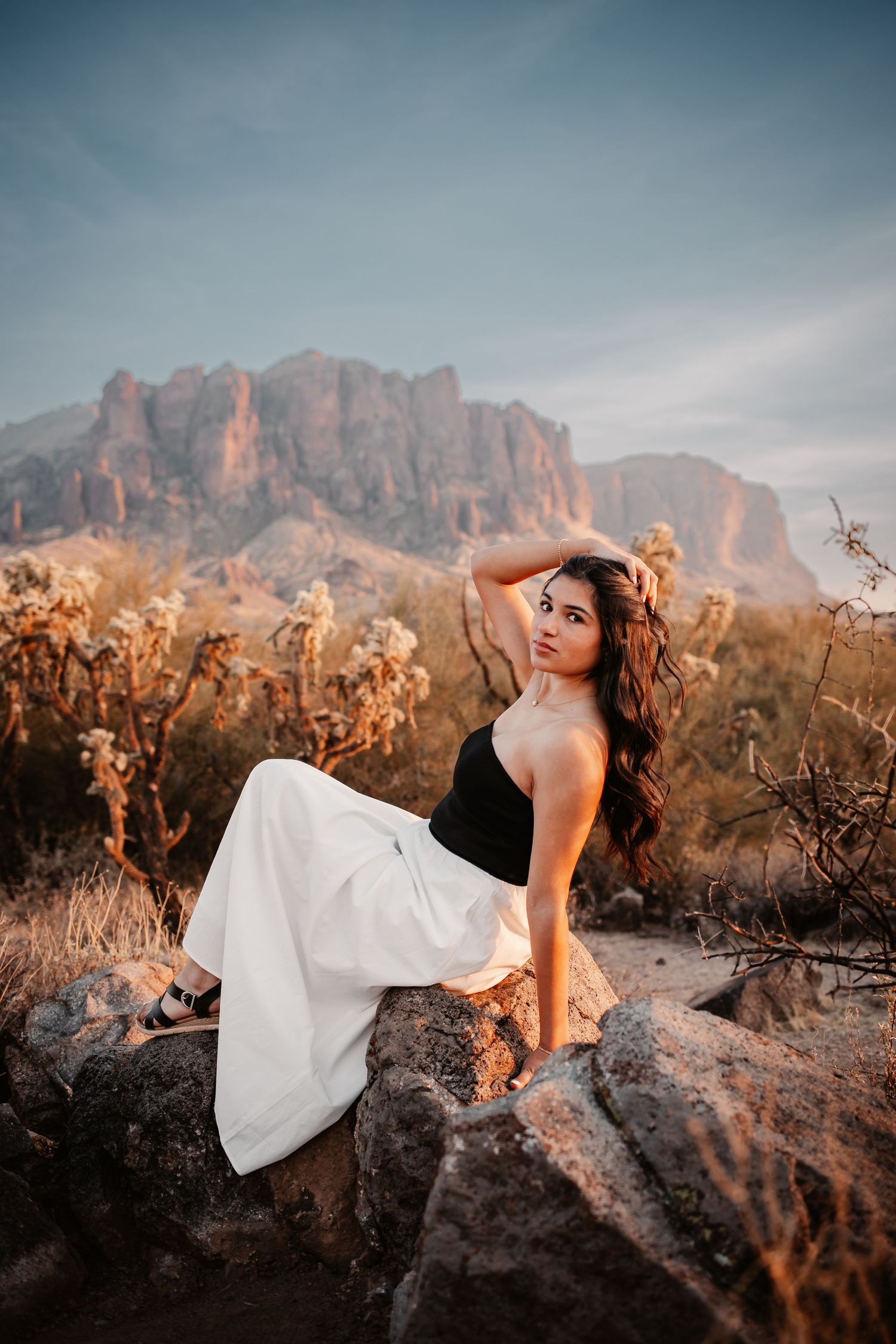 Woman in black top and white pants sits on rocks with Superstition Mountains backdrop at sunset.