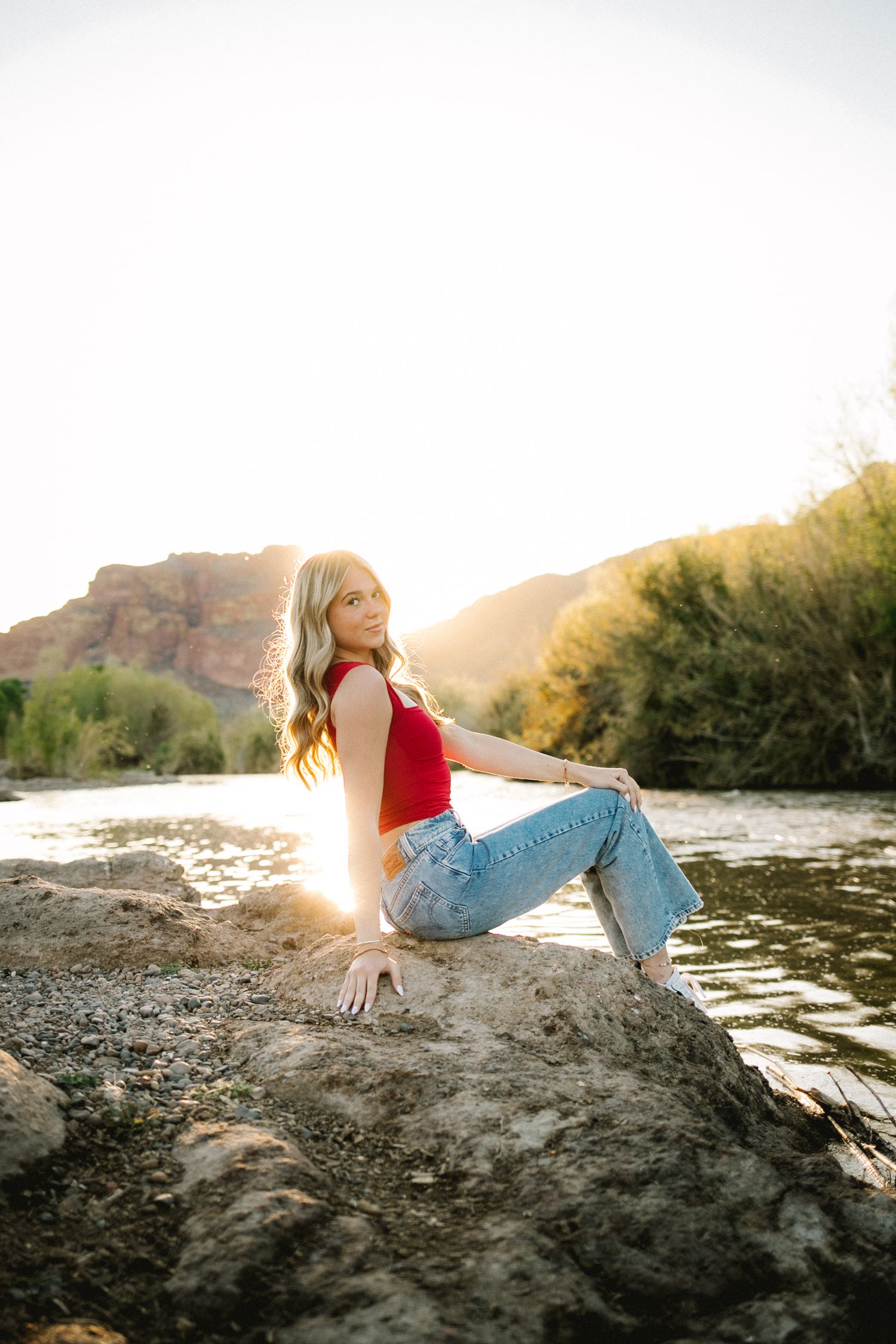 A young blonde woman in a red top and jeans sits on a rock by a river with a sunlit mountain backdrop.