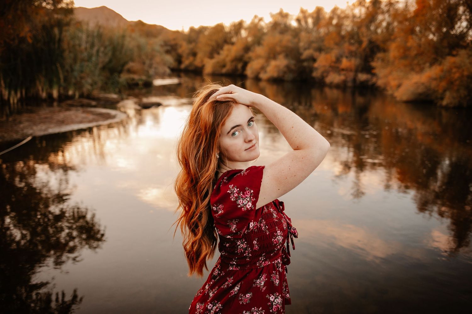 A redhead woman in a floral red dress poses by a reflective autumn lake at golden hour.